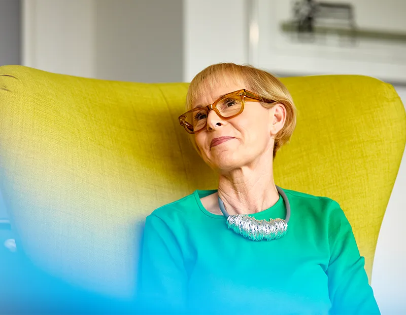 A woman in glasses and a necklace sits in a green wing-backed chair