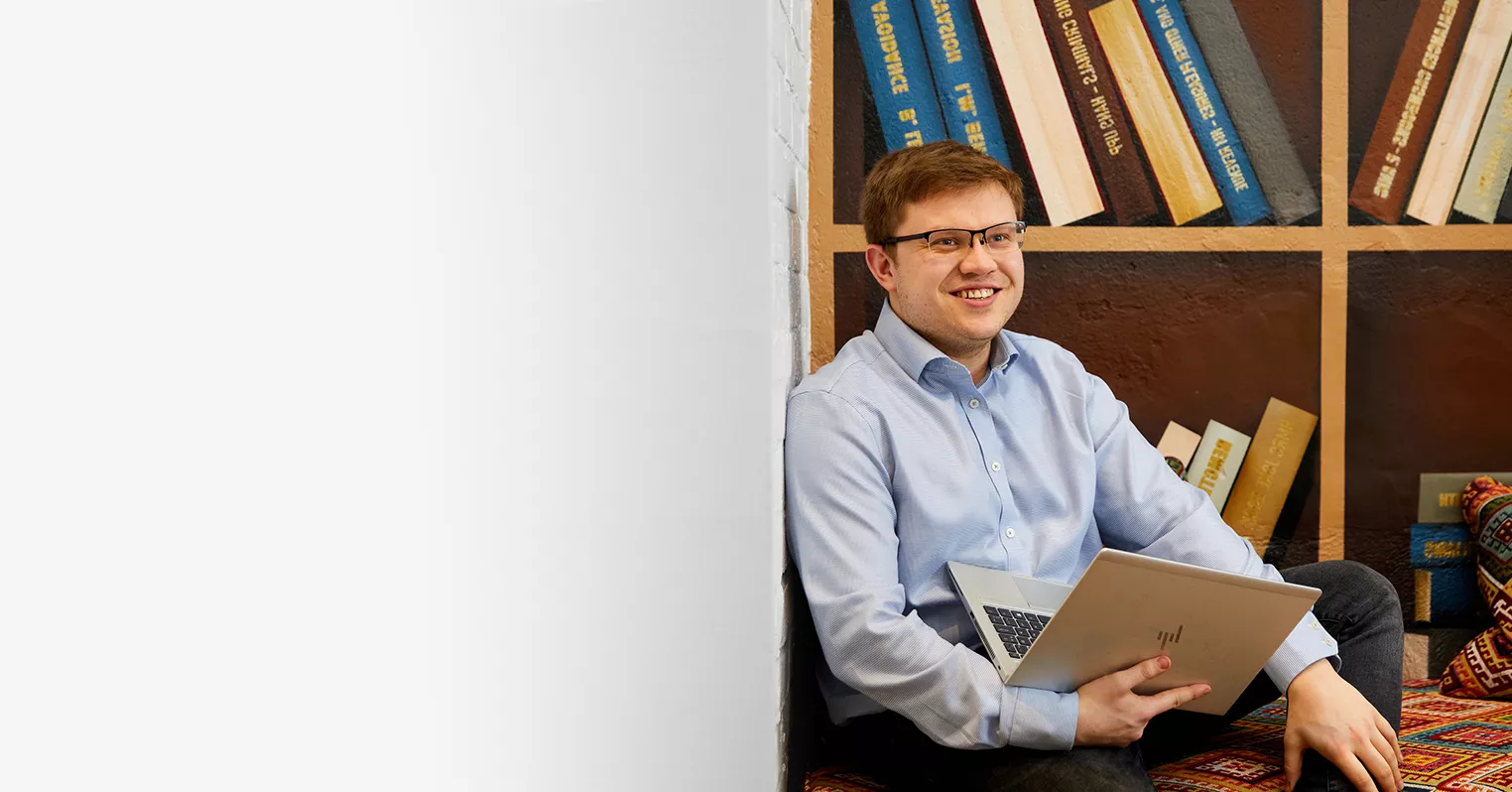 A smiling man sat on a rug holding an open laptop on his lap