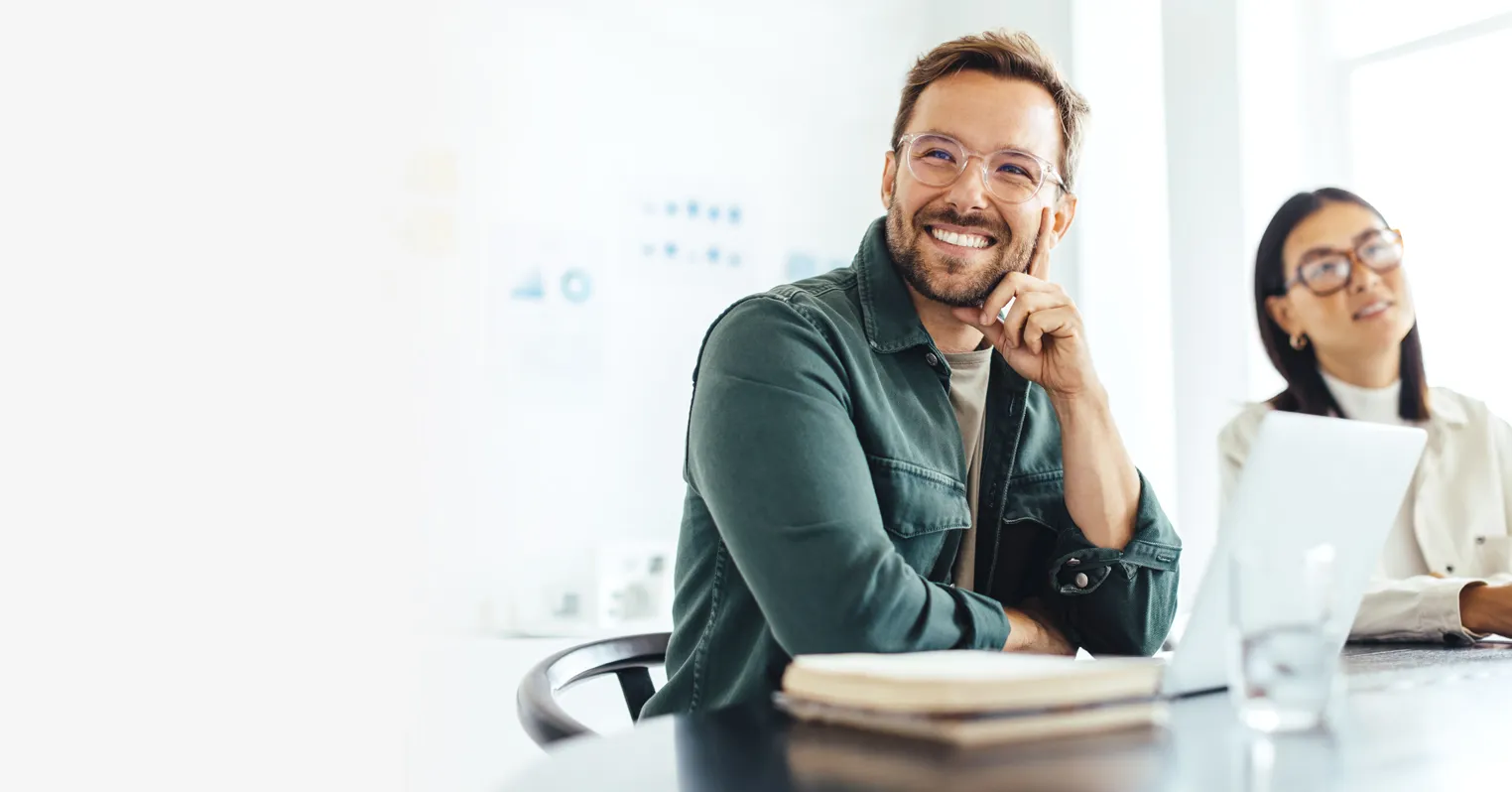 A man wearing glasses sits at a desk and smiles, next to an out of focus woman