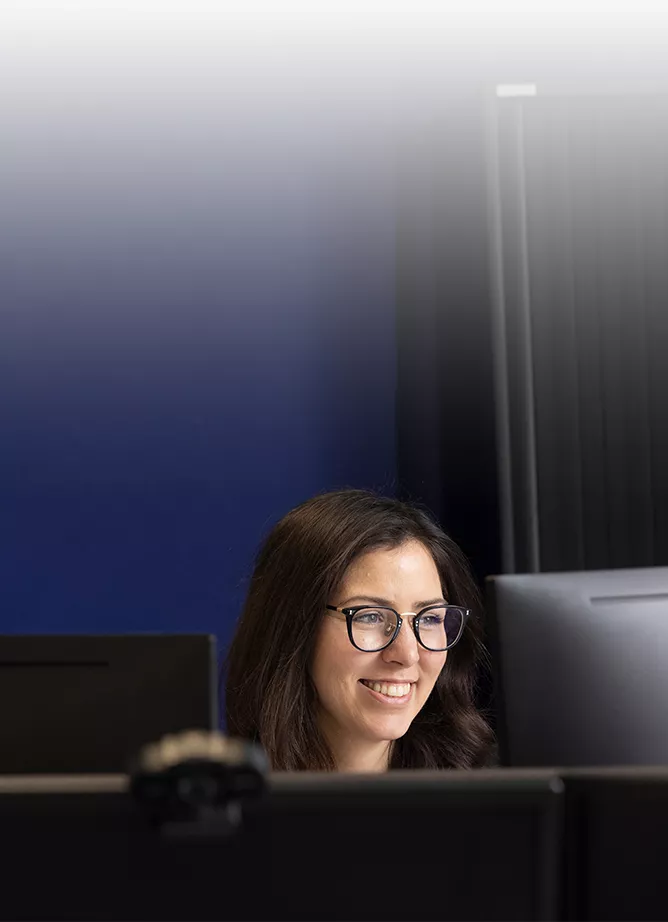 Photo of woman with glasses at desk in front of monitor. Framed wall picture of cartoon dinosaur behind her.